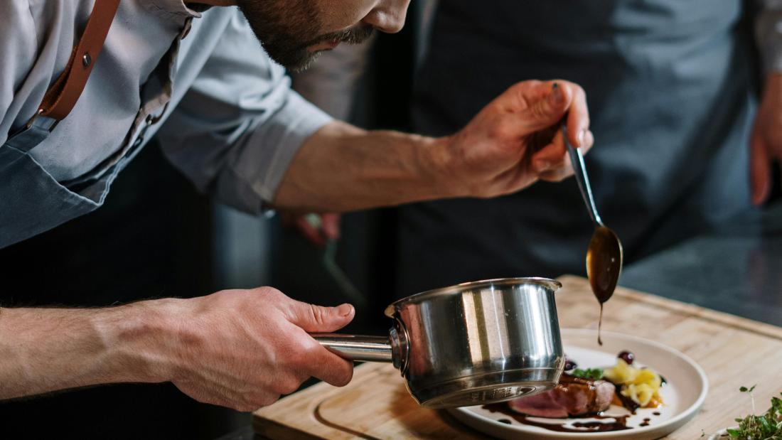 Chef in Blue Shirt Holding Silver Pan adding finishing touches to plate of food