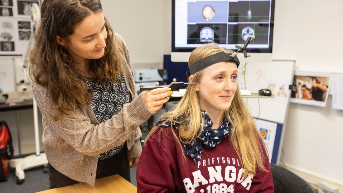 A Psychology lecturer using a neuronavigation pointer to localise a stimulation site on a student’s scalp during preparation for a transcranial magnetic stimulation (TMS) session, with brain images displayed on a computer screen in the background.