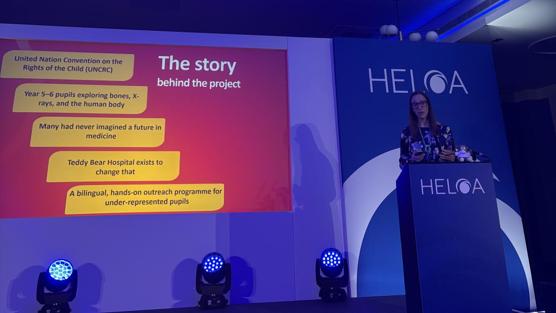 A speaker stands at a podium with the HELOA logo during a presentation. A large screen displays text about an outreach project called Teddy Bear Hospital. The text includes:  “United Nation Convention on the Rights of the Child (UNCRC)” “Year 5–6 pupils exploring bones, X-rays, and the human body” “Many had never imagined a future in medicine” “Teddy Bear Hospital exists to change that” “A bilingual, hands-on outreach programme for under-represented pupils.” Stage lighting and a blue backdrop are visible.