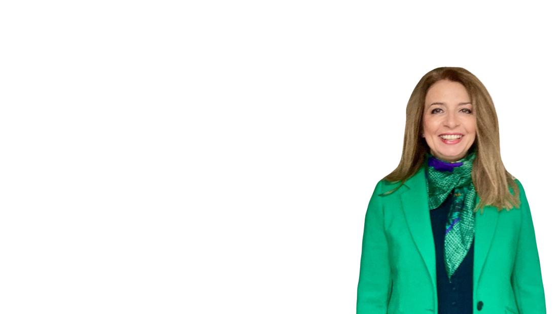 Person standing against a plain white background, wearing a bright green blazer over a dark top with a patterned green scarf