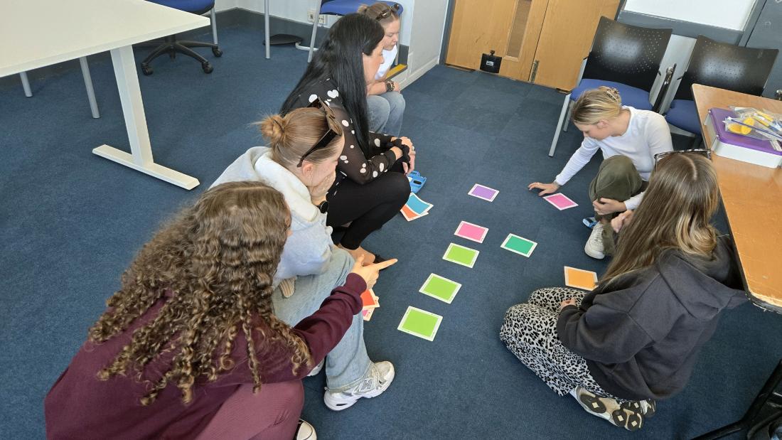 Group of people sitting on a carpeted classroom floor, arranging and pointing at coloured square cards placed in a grid during a collaborative activity
