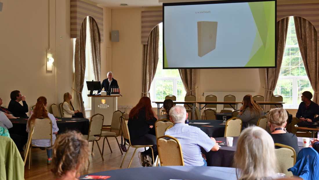 Participants seated at tables in a large event hall watch a presentation being delivered from a lectern at the front of the room. A presentation slide is displayed on a large screen.
