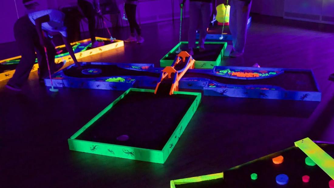 Four courses of UV mini golf in the dark, with players surrounding the courses with putters and golf balls.
