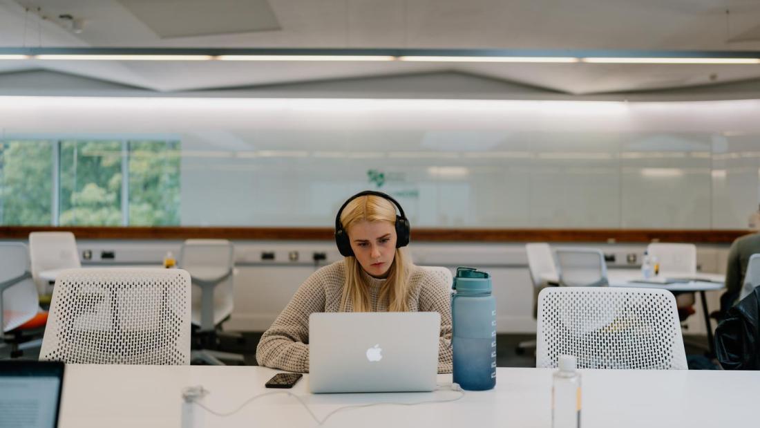 A student wearing headphones is sat at a white table working on a laptop in the Social Learning Space at Pontio