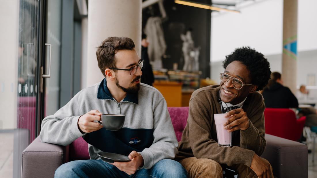 Two students socialising in a café, one is holding a hot drink and another is holding a smoothie.