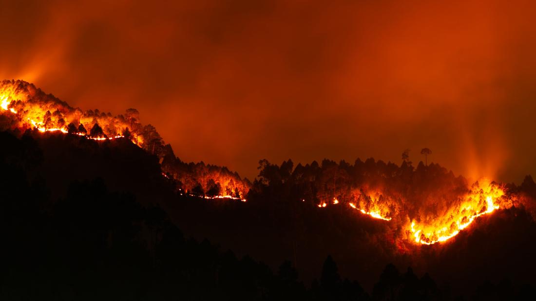 A blazing forest fire spreads across a mountainous landscape, illuminating the night sky