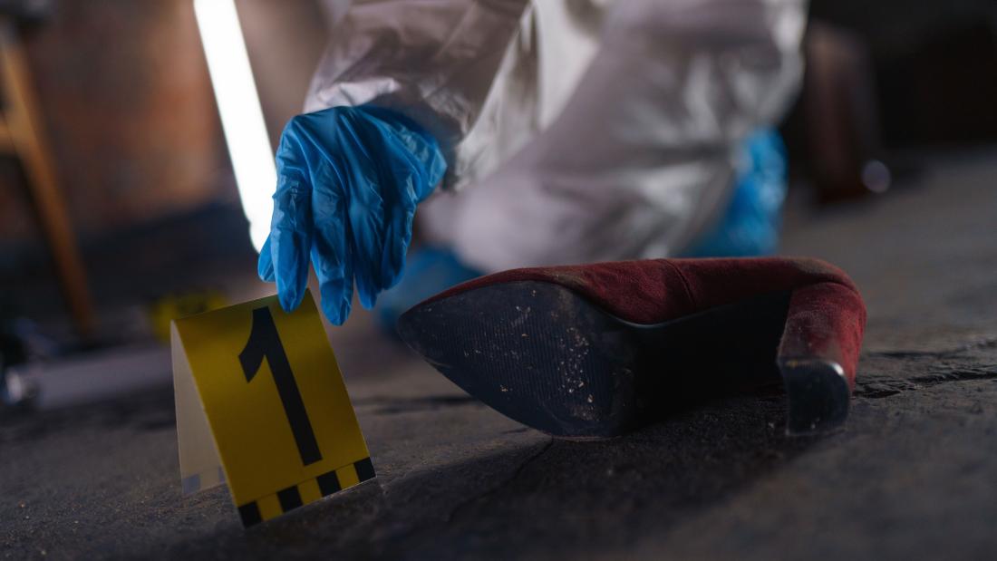 A forensic investigator wearing protective clothing and blue gloves places an evidence marker beside a red high-heeled shoe at a crime scene.