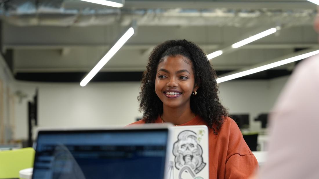 Student smiling sat in front of a laptop in the colab space in Pontio