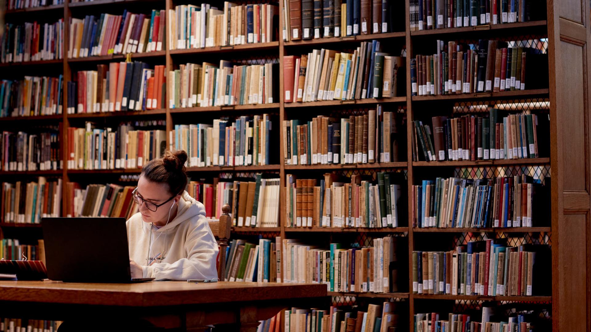 Student sitting in the library with a laptop and books on shelves in the background