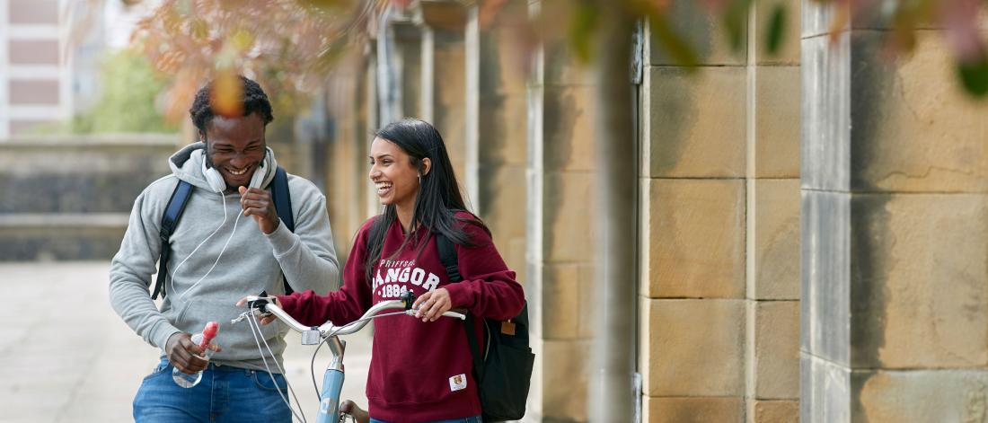 Two students walking and pushing a bike outside the Main Arts building