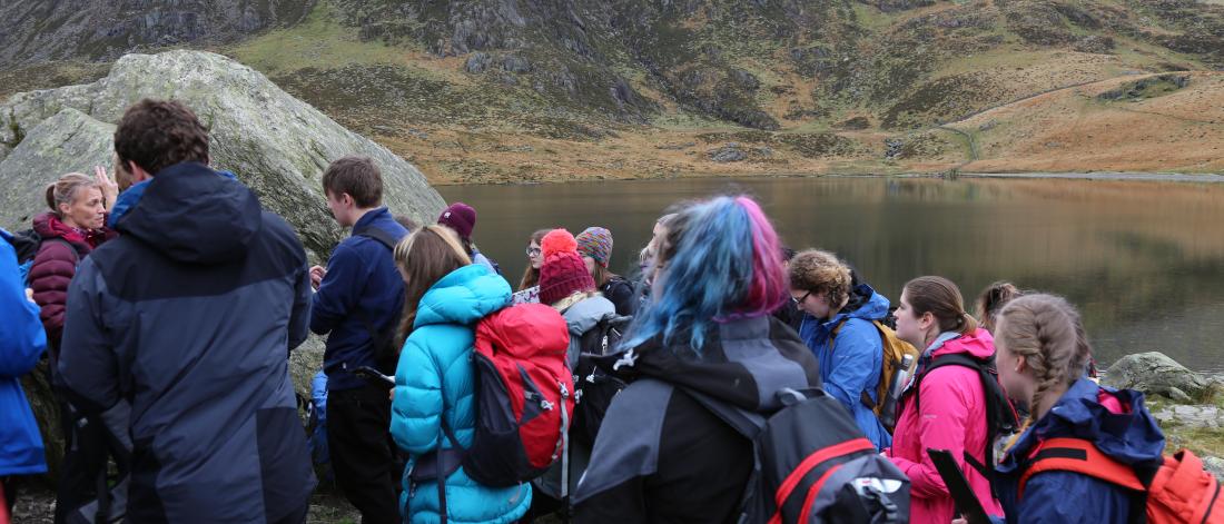 A group of students standing next to a lake, listening to a lecturer talking - on a field course in Cwm Idwal