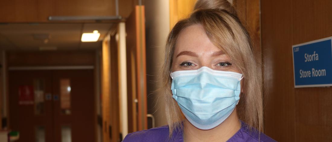 A student nurse wearing  purple uniform and a mask looks into the camera