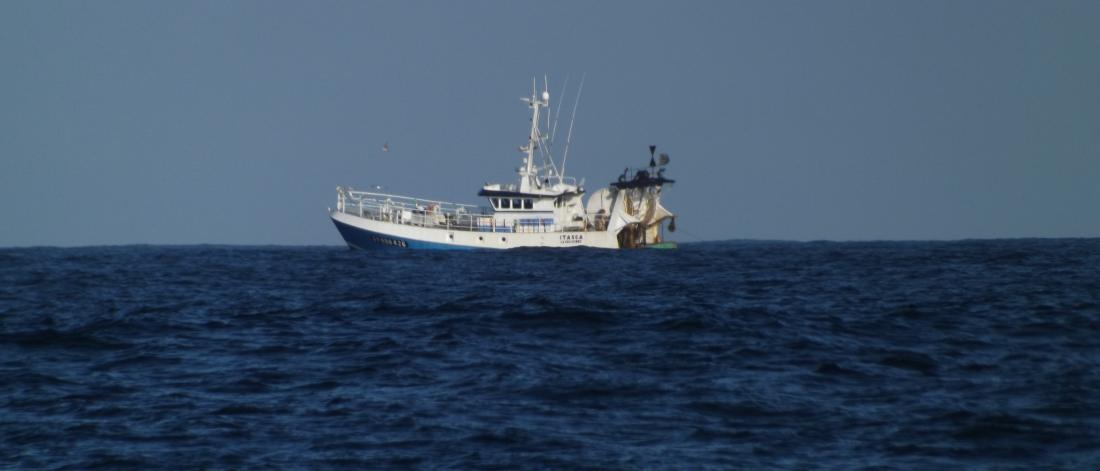 A blue trawler sits near the horizon on a blue sea under a lighter blue sky.