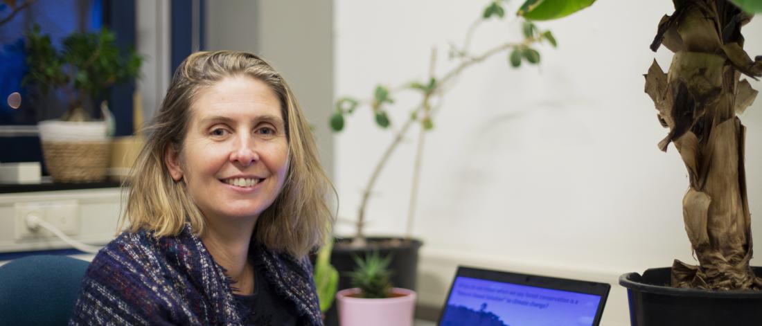 Professot Julia Jones, with blonde shoulder length hair smiles into camera, she is seated in front of a laptop computer and large tropical looking houseplant.