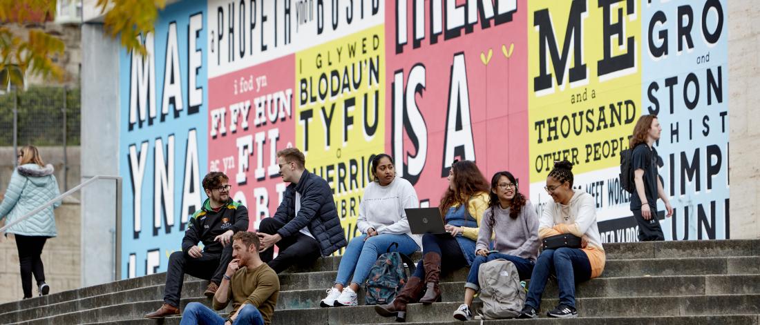 Bangor University students sitting on a stairs with graffiti in the background