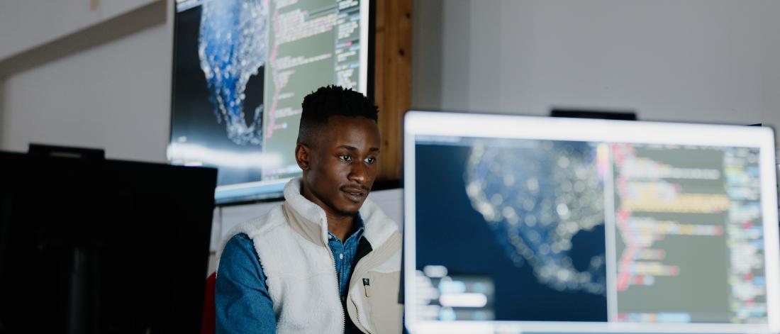 A student sitting in front of a computer with a large screen in the background and a smaller screen in the foreground  