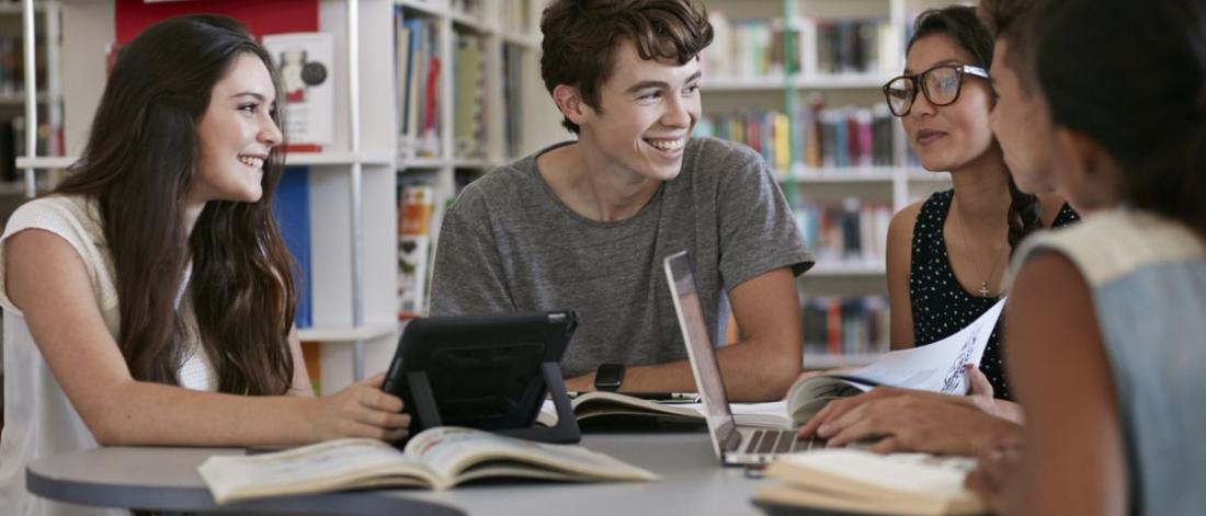 Students sitting at a table talking. On the table are laptops and books.