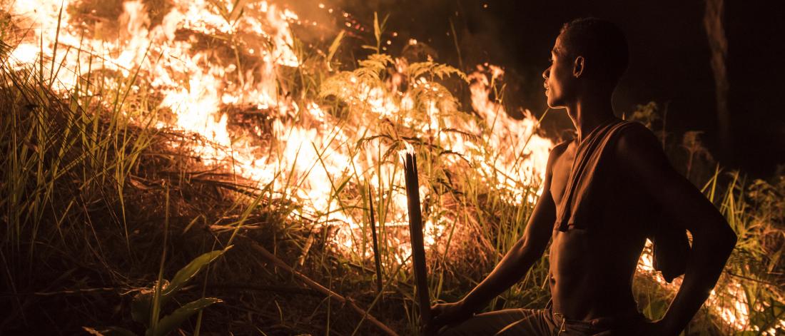 A man stood by some burning vegetation, he is lite by the bright orange flames of the fire.