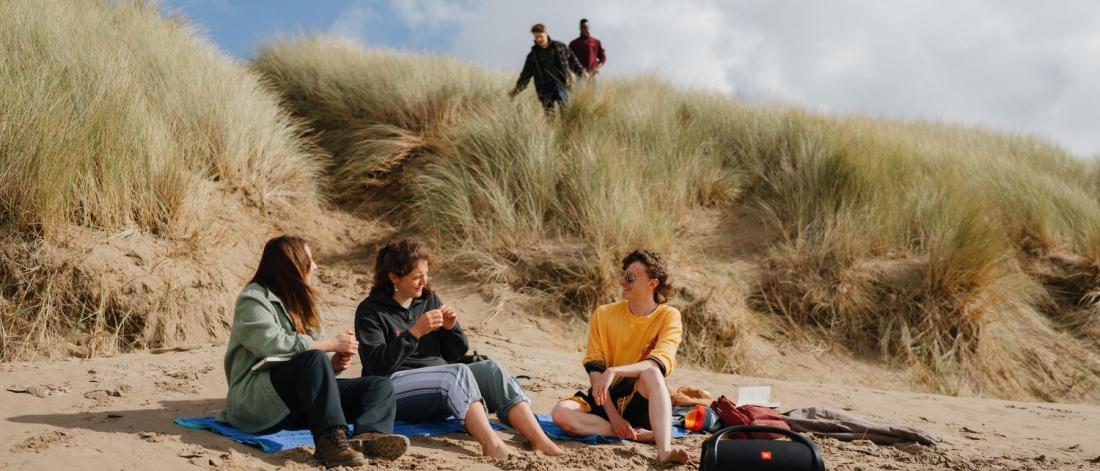 Three students sitting on the sand chatting and two students are walking towards them through the dunes behind them