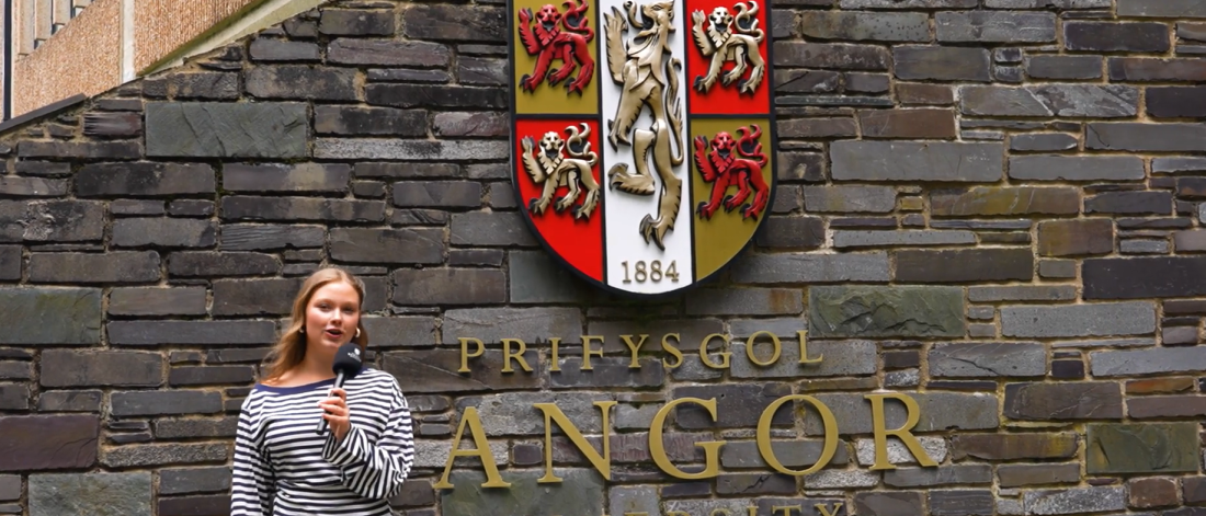 Myfyriwr yn gafael mewn microffôn yn sefyll o flaen wal gyda logo Prifysgol Bangor arno