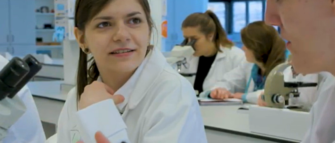 Students wearing white lab coats sitting chatting and looking into microscopes at a laboratory