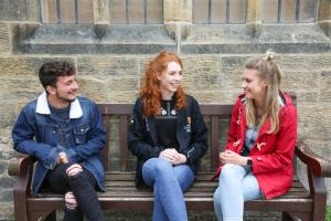 Three students sat on a bench on campus talking to each other 