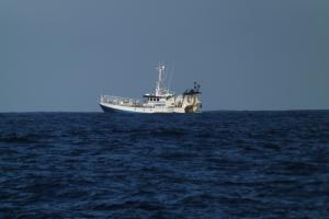 A blue trawler sits near the horizon on a blue sea under a lighter blue sky.