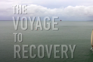 The Voyage to Recovery video screen shot- these words appear to the left of a shot looking down as a sailing boat and some coastline