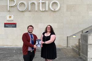 Dau fyfyriwr o Brifysgol Bangor yn dangos eu cyhoeddiad cyntaf y tu allan i adeilad Pontionts show the book they have published while standing in front f the external of the Pontio building