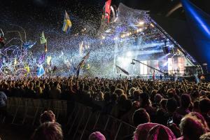A large crowd looks towards a brightly lit stage at night. Above their heads are coloured banners.