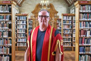 A middle aged man in spectacles and red academic gown stands in the beutiful Shankland Library, Bangor University