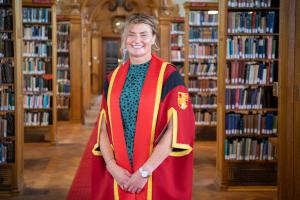 Toung woman smiles into camera in library setting, wearing red graduation gown