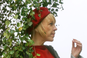 Woman with blonde hair wearing red and a red beret stands directly under a tree in leaf.