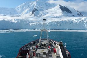 The bow of a ship is seen in front of  snow covered mountains and a glacier running down to a blue sea.