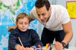 Young man standing with a younger male child who is sitting at a desk