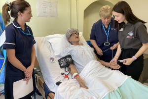 YOung girl approached a dummy in a bed- there are nurses on either side.