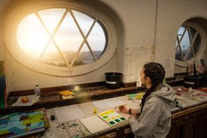 A student with plaits at a work desk, looks through a window