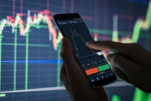 someone pointing towards their phone with finical stock market information on the phone and on screen in background