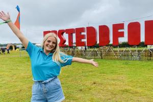 A girl with long blonde hair, wearing a Bangor turquoise t shirt raises her arms, behind her are large red letters spelling EISTEDDFOD