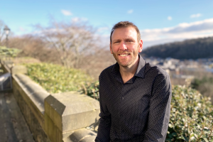 Person smiling whilst leaning against a stone wall with urban landscape in the background