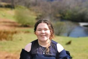 Menyw â gwallt brown wedi’i glymu’n ôl yn gwisgo ffrog glas tywyll yn sefyll yn gwenu yn Eryri. Mae coed, mynyddoedd a llyn i’w gweld yn y cefndir. 