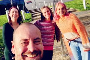 Image of 4 members of the Annedd Ni/Blas Pontio team outside Pontio building in Bangor. The image shows a man in a black t-shirt smiling, behind him are 3 women wearing green, pink and orange. In the background, you can see the steps leading up to Pontio and the Pontio sign