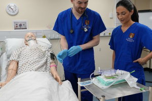 a simulation mannequin is lying in a hospital bed in a university clinical skills suite. two medical students a male and a female are stood to the side with a trolley