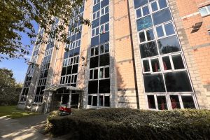 Tall building in striking pinkish brick. lots of windows and stylish black panelling between windows. Surrounded by greenery.