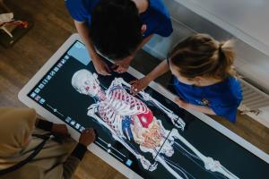a human skeleton is shown on the screen of a digital anatomy table from above with 2 students and a lecturer stood around it