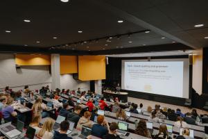 A view of the lecture theatre from the back of the room looking down on a room full of students looking at the projector screen at the front of the room.