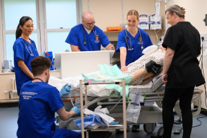 4 medical students dressed in blue scrubs stand around a mannikin in a hospital bed, there is an intructor in a black tunic overseeing the students