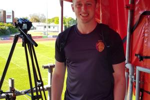 George Lowe in rugby tshirt, smiling at camera, with a tripod and a green pitch behind him 