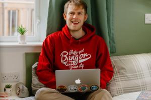 Male student wearing a Bangor University red branded hoodie sat on a single bed with a  laptop on his lap