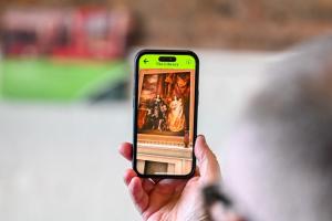 Photograph of a hand holding a smart phone, displaying the Margam app. 